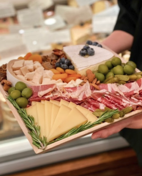 Image of a small custom cheese and charcuterie board made up of wedges, chipped, and quarters of cheeses and thinly sliced cured meats surrounded by olives, fresh and dried fruits, and nuts being held in front of the cheese case.